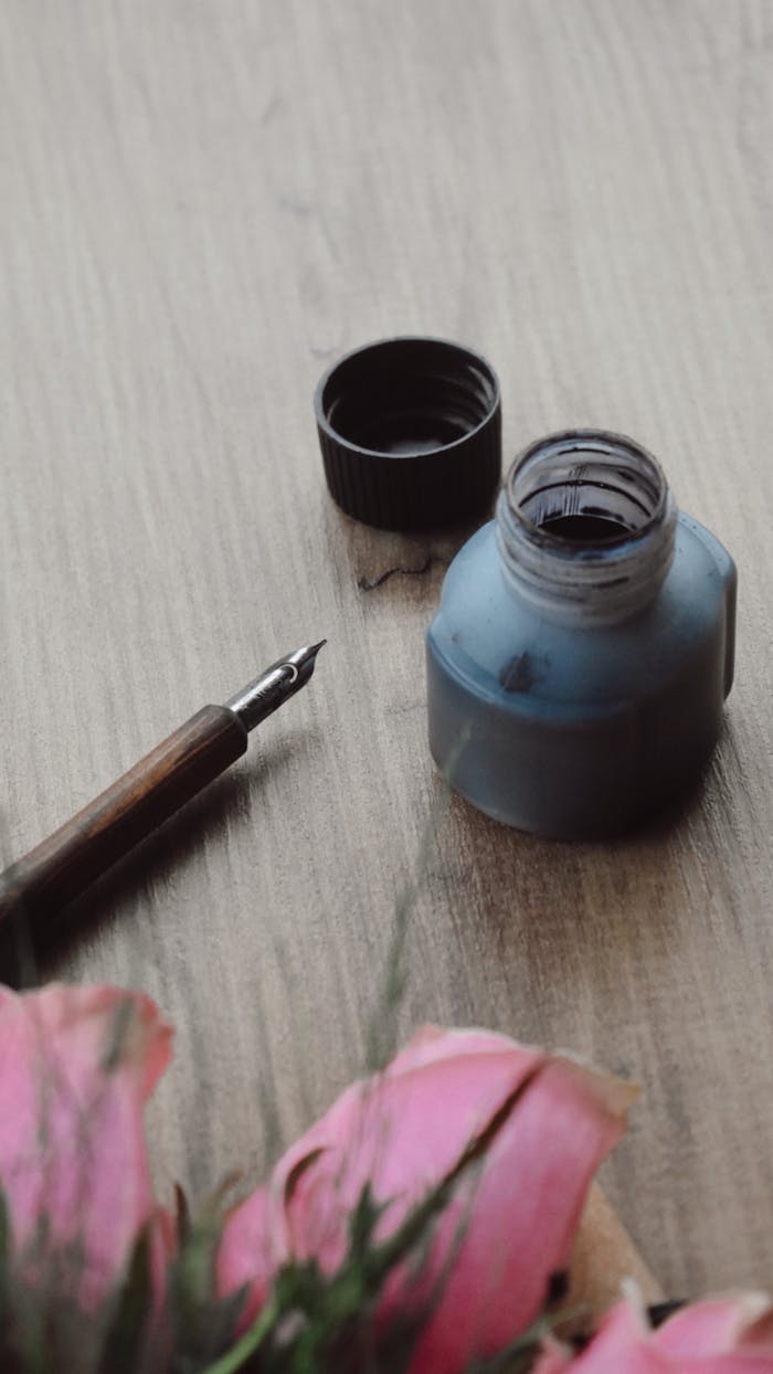 Close-up of a fountain pen with ink bottle and pink flowers on wooden surface.