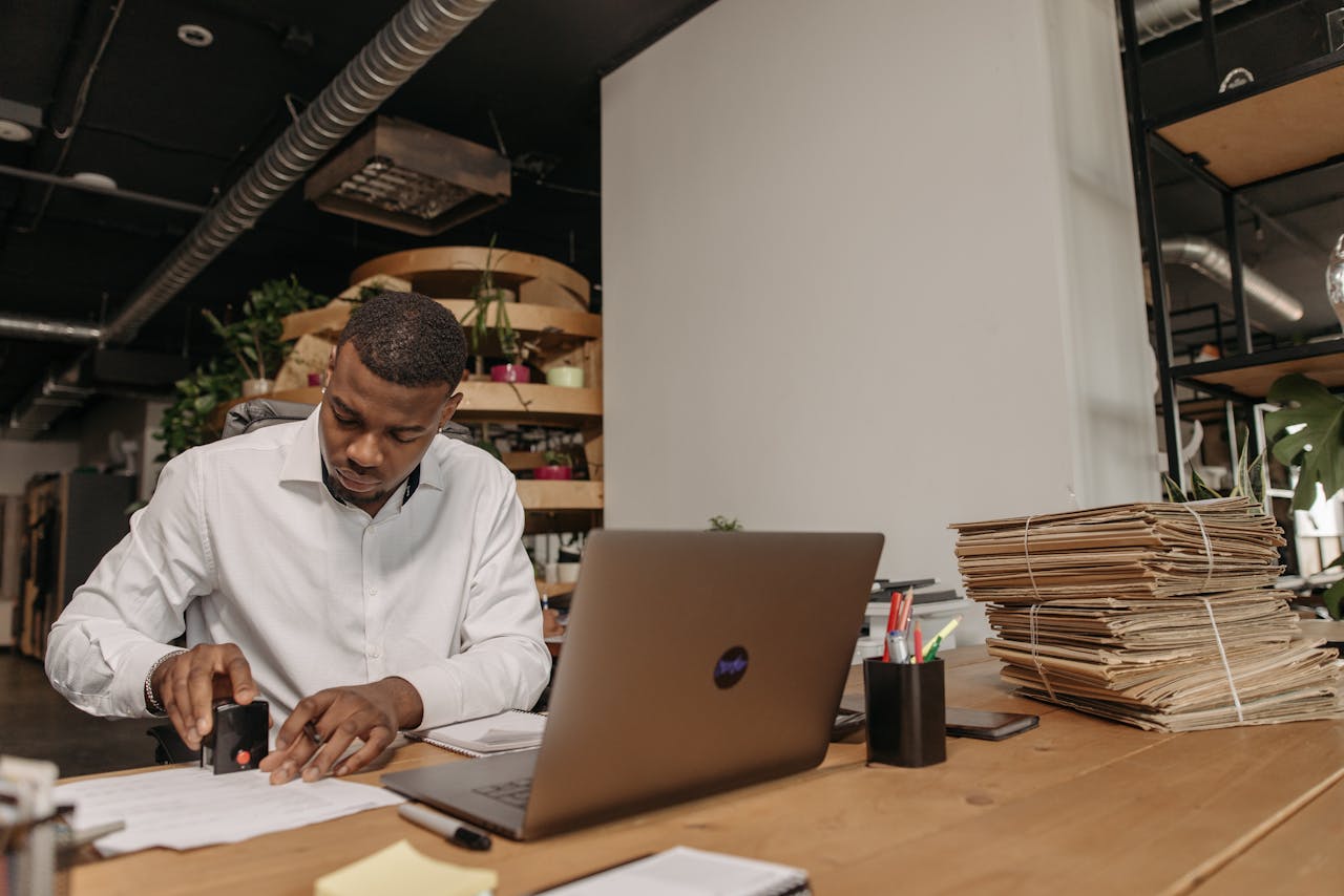 African American man focused on stamping documents at an office desk with a laptop and folders.