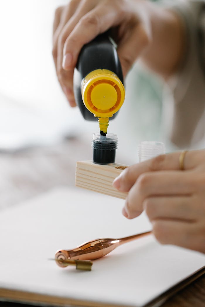 A hand carefully pouring black ink into a small container with calligraphy tools nearby.